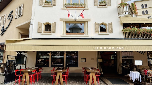 Ein Restaurant mit Außenbereich, einem Schild mit der Aufschrift "LE HIBOU BLANC" und einem Gebäude mit mehreren Fenstern und Balkonen.