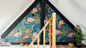 A room with a sloped ceiling, a wooden staircase, a wall with a flamingo wallpaper, a potted plant, and a small table with books and candles.