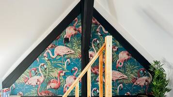 Een kamer met een schuine plafond, een houten trap, een behang met flamingo’s, een potplant en een kleine tafel met boeken en kaarsen.