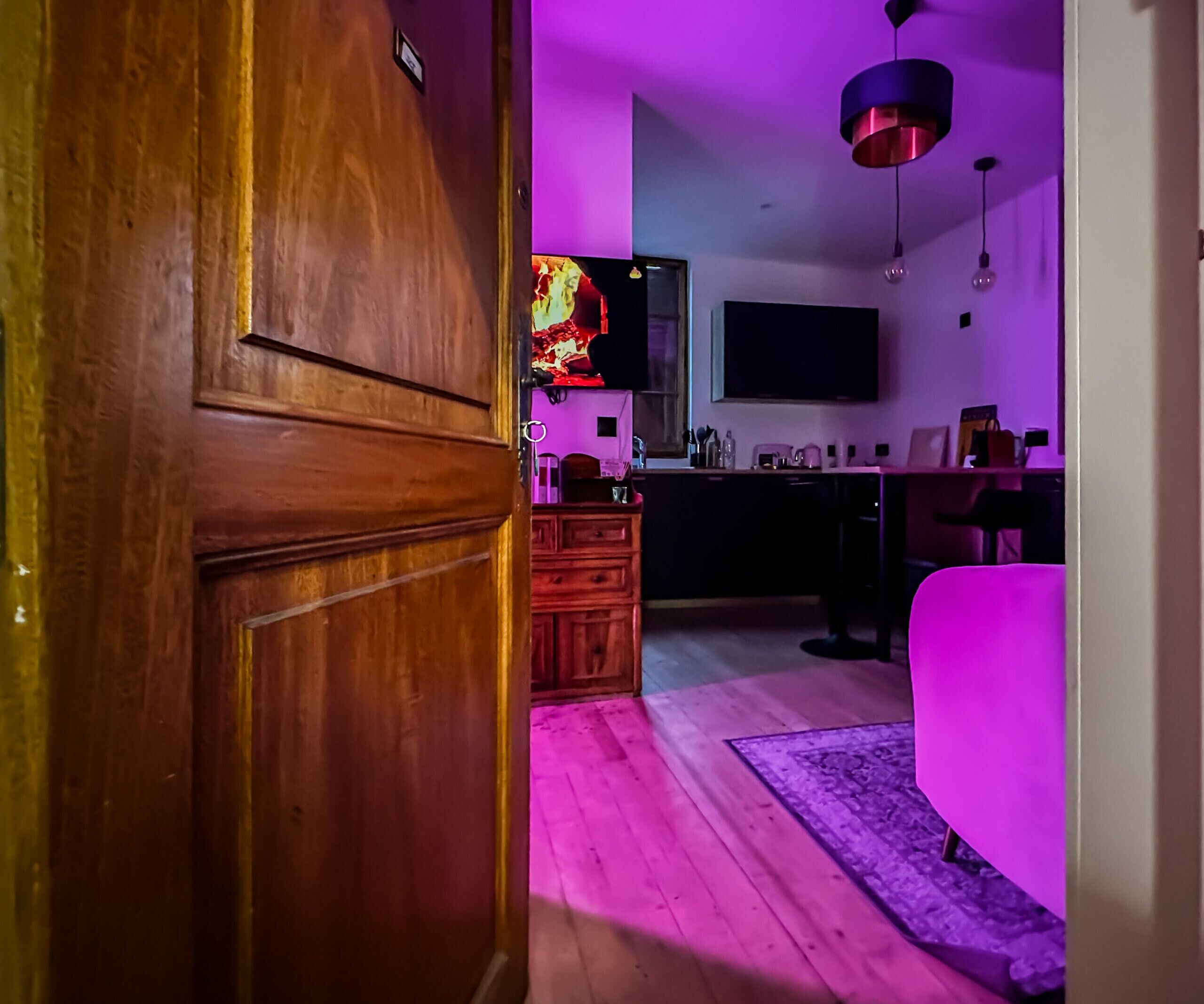 Een kamer met een houten deur, een keukenhoek met een televisie, een eettafel en een roze stoel.