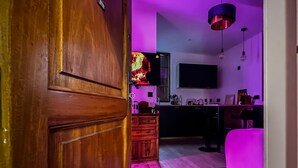 A room with a wooden door, a kitchen area with a TV, a dining table, and a pink chair.