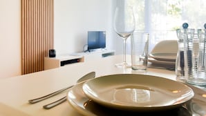 Mesa de jantar posta com pratos, talheres e copos, com uma televisão e janela ao fundo.