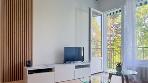 Uma sala de estar moderna com televisão em um armário branco, uma mesa de vidro e vista para o verde lá fora.