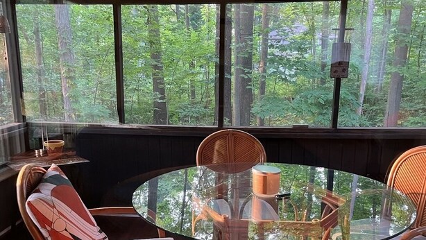Ein runder Glastisch mit vier Korbstühlen, umgeben von einer Holzterrasse mit Blick auf einen Wald.