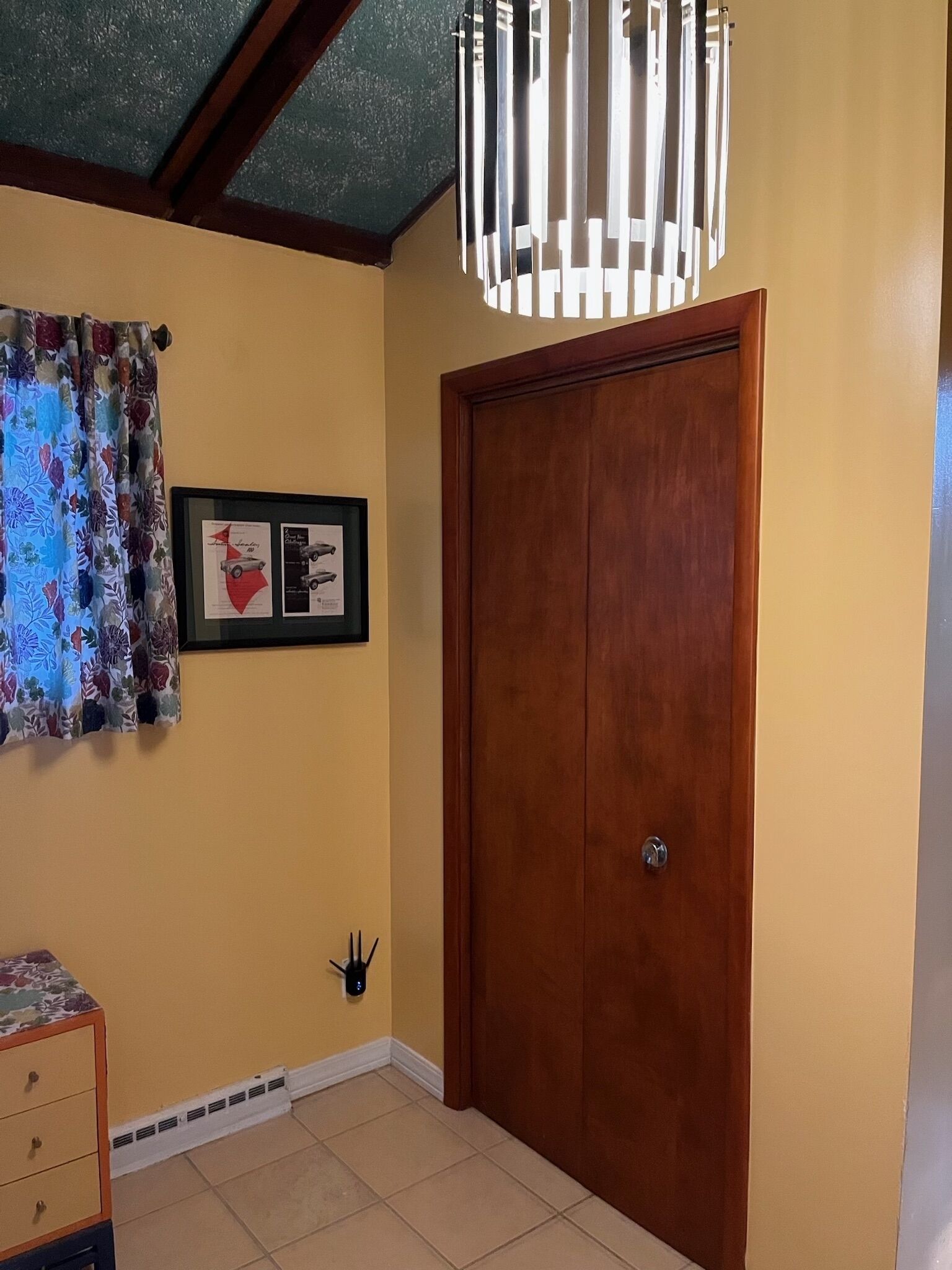 A room with a wooden door, a framed picture on the wall, a patterned curtain, and a small table with drawers.