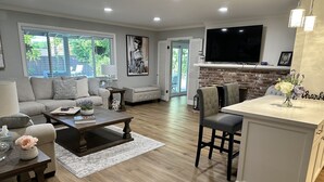 A modern living room with a fireplace, a dining area, and a kitchen with a large TV. - Secluded Home w/pool located 5 mins from American River  (Carmichael)