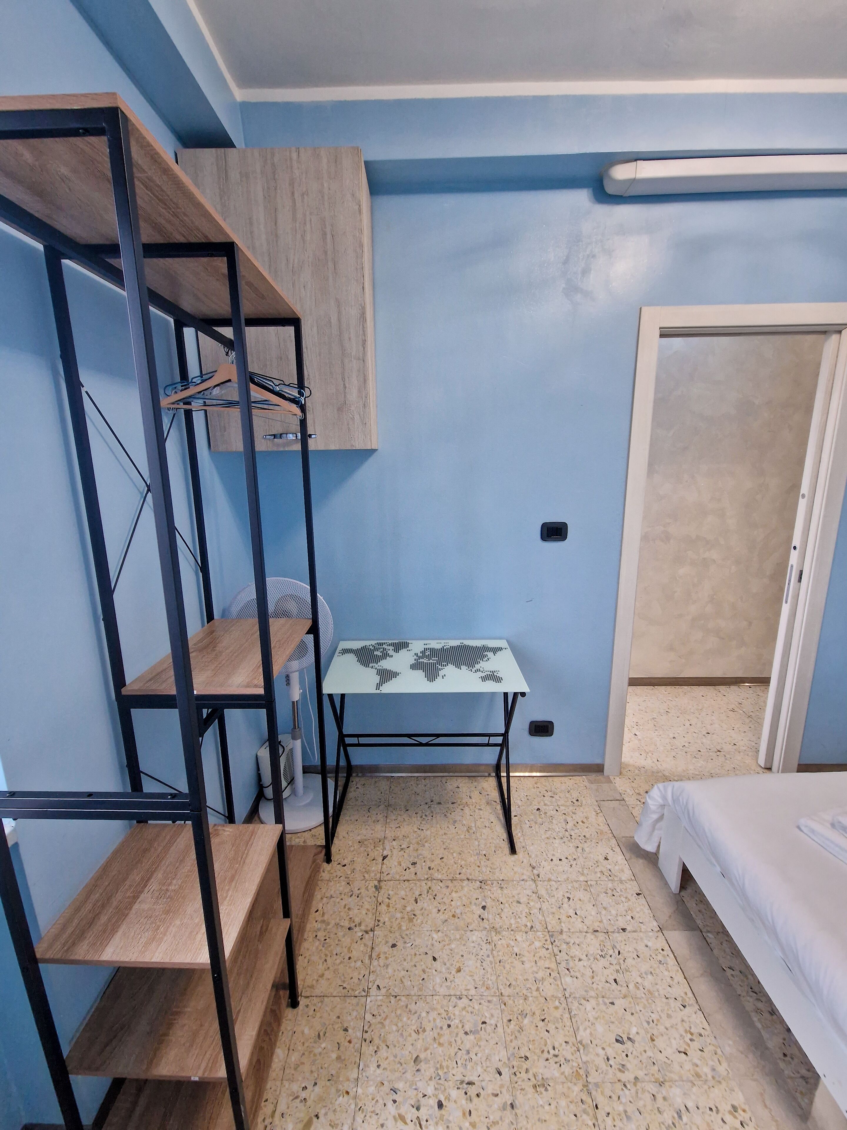 Ein kleines Zimmer mit Etagenbett, einem kleinen Tisch mit Weltkarten-Motiv, einer Holzregaleinheit und einem Ventilator.