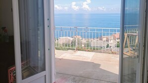 A room with a large sliding glass door leading to a balcony with a view of the sea and houses.