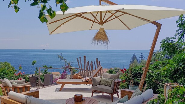 An outdoor seating area with a view of the sea, wooden furniture, and a large umbrella.