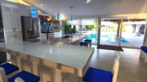 A modern kitchen with a large island, a dining area with blue chairs, and a pool area visible through glass doors. - Villa Kassondra del Mar (Acajutla)