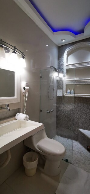 A modern bathroom with a white toilet, a sink with a mirror, a hairdryer, and a shower with a glass enclosure. - Villa Kassondra del Mar  (Acajutla)