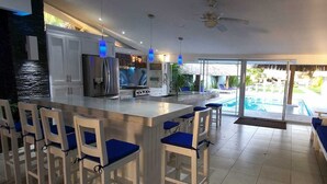 A modern kitchen with a large island, bar seating, and a view of a swimming pool. - Villa Kassondra del Mar (Acajutla)