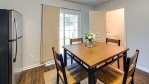 Un comedor con mesa de madera, cuatro sillas, un jarrón con flores, un refrigerador y una ventana con cortinas.