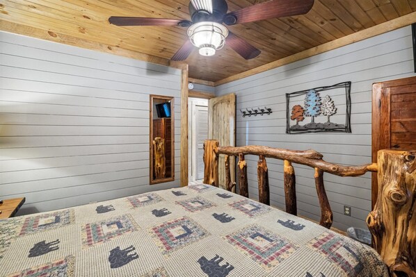 Een slaapkamer met een plafondventilator van hout, een bed met een quilt met een patroon, een houten hoofdbord, en een wandkapstok met een decoratie in boomvorm.
