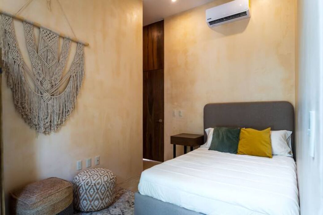 Quarto com cama, um painel de macramé e uma pequena mesa de cabeceira.