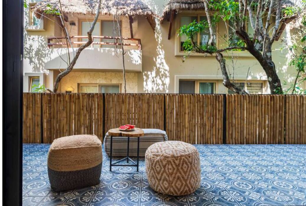 A patio with a patterned floor, two ottomans, a small round table with a red object, and a bamboo fence.