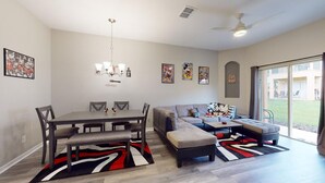A modern living room with a dining table, sofa, and ottoman, featuring artwork and a large sliding glass door to the outdoors. - Themed, 4 Bed, 3bth, Next To Pool, Water Slides, Walk To Oldtown, 6 to Disney. (Kissimmee)
