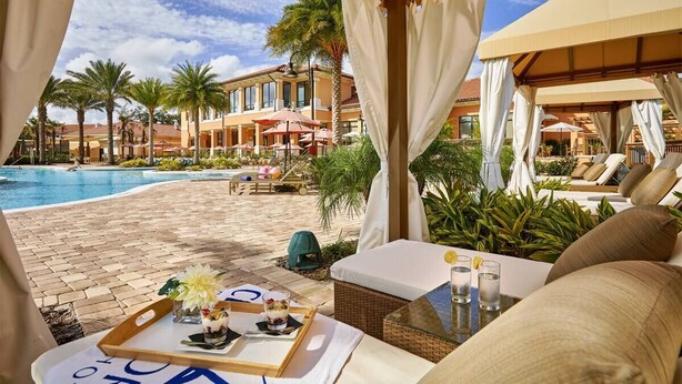 A poolside area with lounge chairs, a dining table with a tray of food, and a view of the pool and buildings in the background. - Themed, 4 Bed, 3bth, Next To Pool, Water Slides, Walk To Oldtown, 6 to Disney. (Kissimmee)