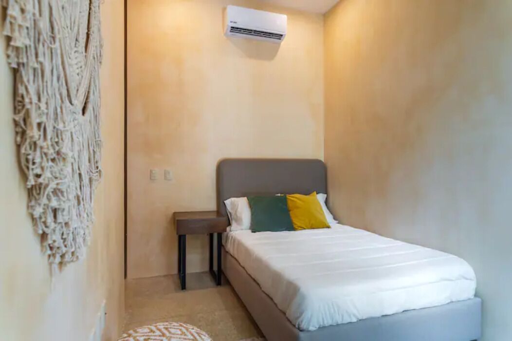 A bedroom with a bed, a bedside table, a wall-mounted air conditioner, and a macramé wall hanging.
