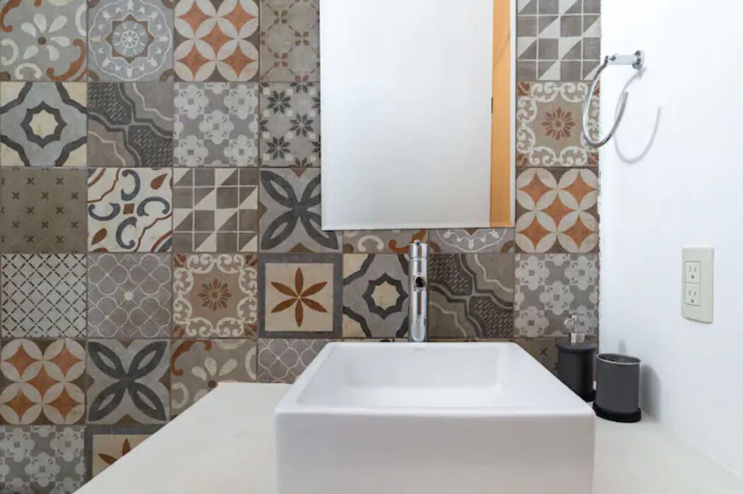 A bathroom with a white sink, a mirror, and a shower. The wall is tiled with various geometric patterns.