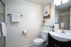 Une salle de bain avec des toilettes, un lavabo et une douche. Un tableau encadré est accroché au mur et un flacon de savon est posé sur le lavabo.