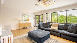 Un salon moderne doté d’une grande fenêtre, d’un canapé foncé, d’un pouf assorti et d’un meuble en bois avec un miroir.