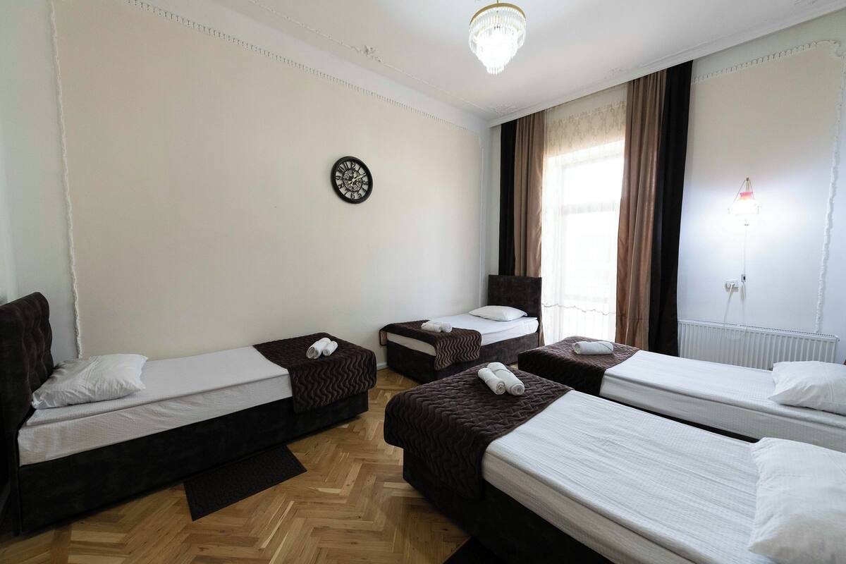 A hotel room with three single beds, a wooden parquet floor, a chandelier, a clock, and a window with curtains.
