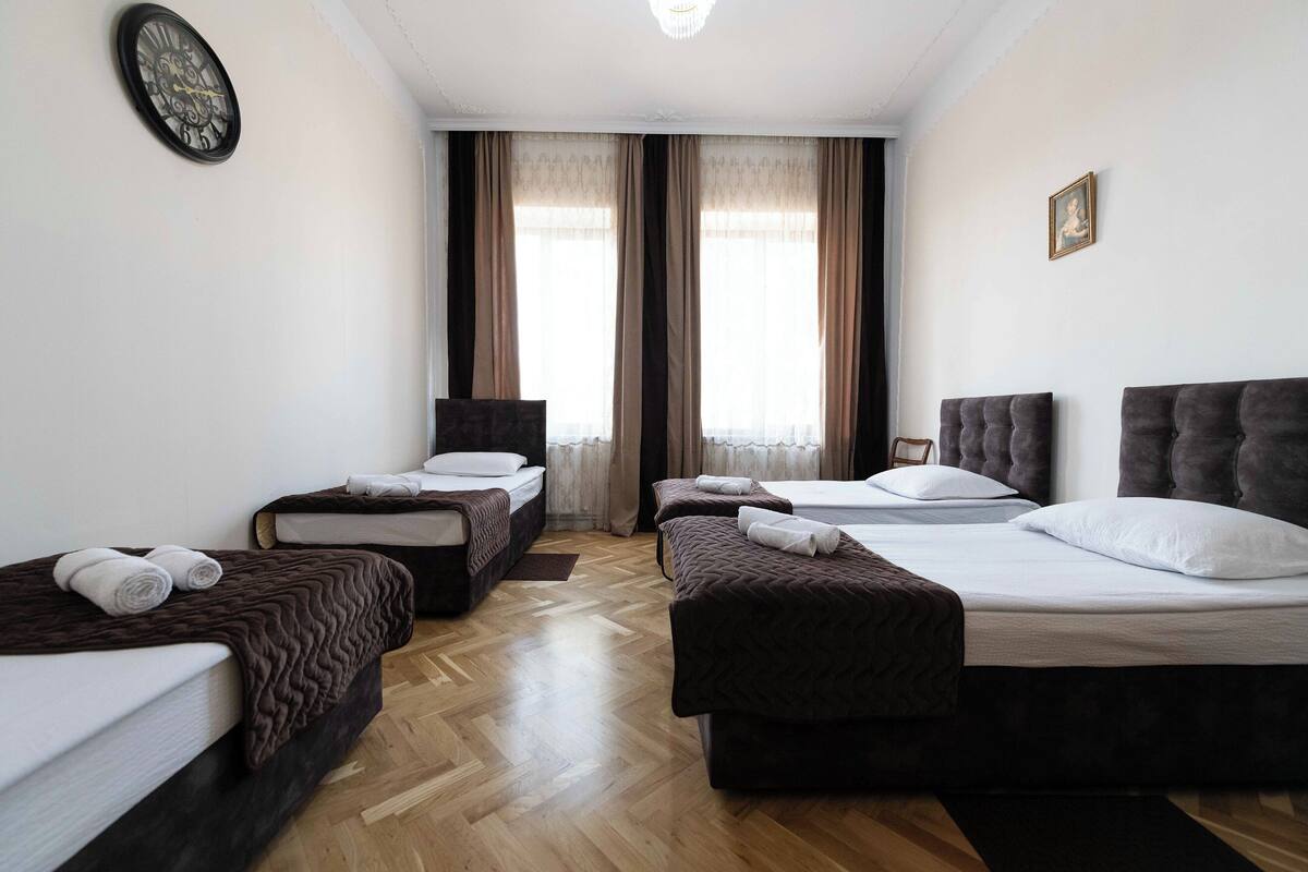 A hotel room with two single beds, a sofa bed, a wooden floor, a clock, and a window with curtains.