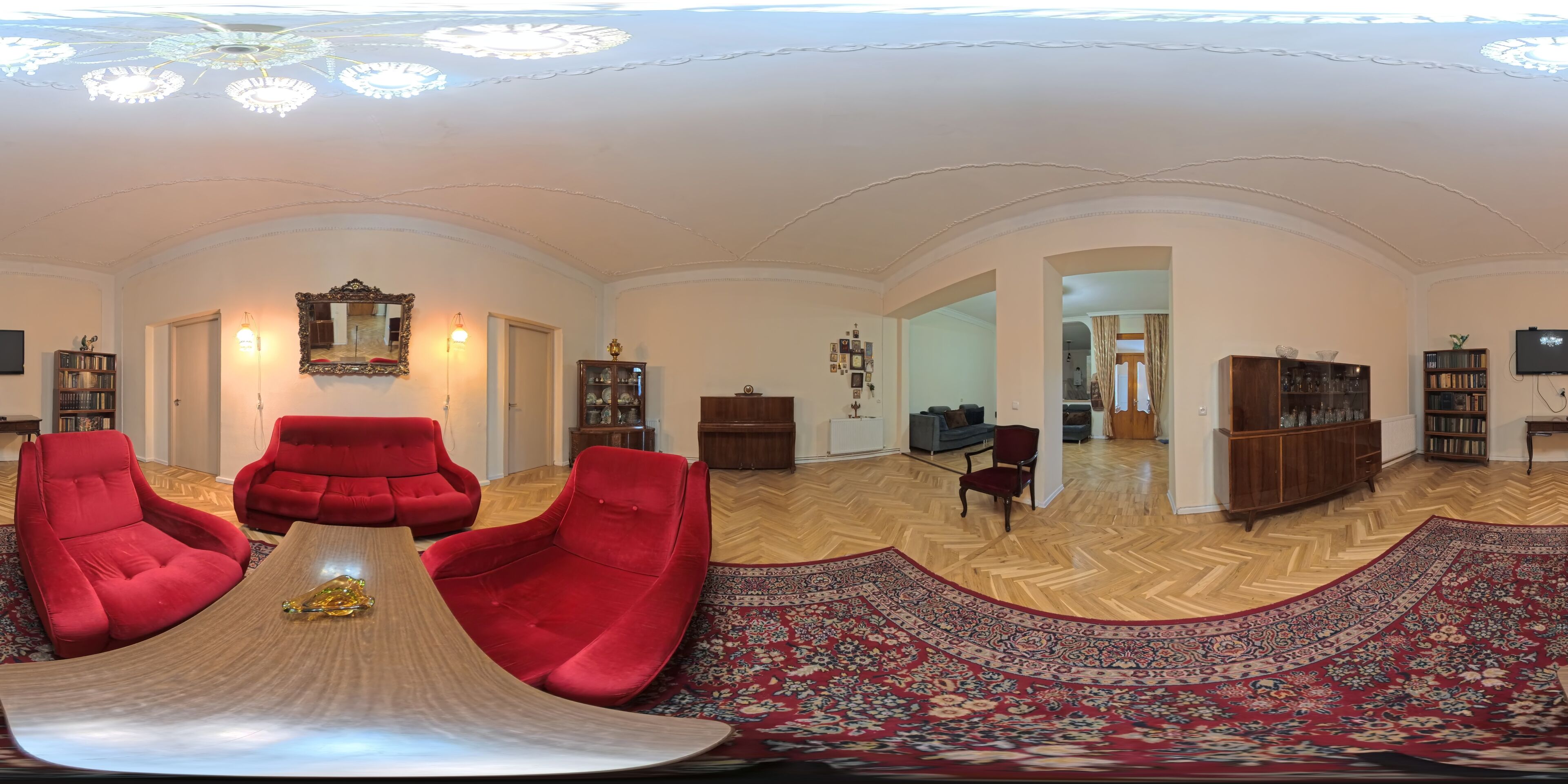 Een ruime woonkamer met een visgraatvloer, rode fauteuils, een houten bijzettafeltje en een groot tapijt met een patroon.