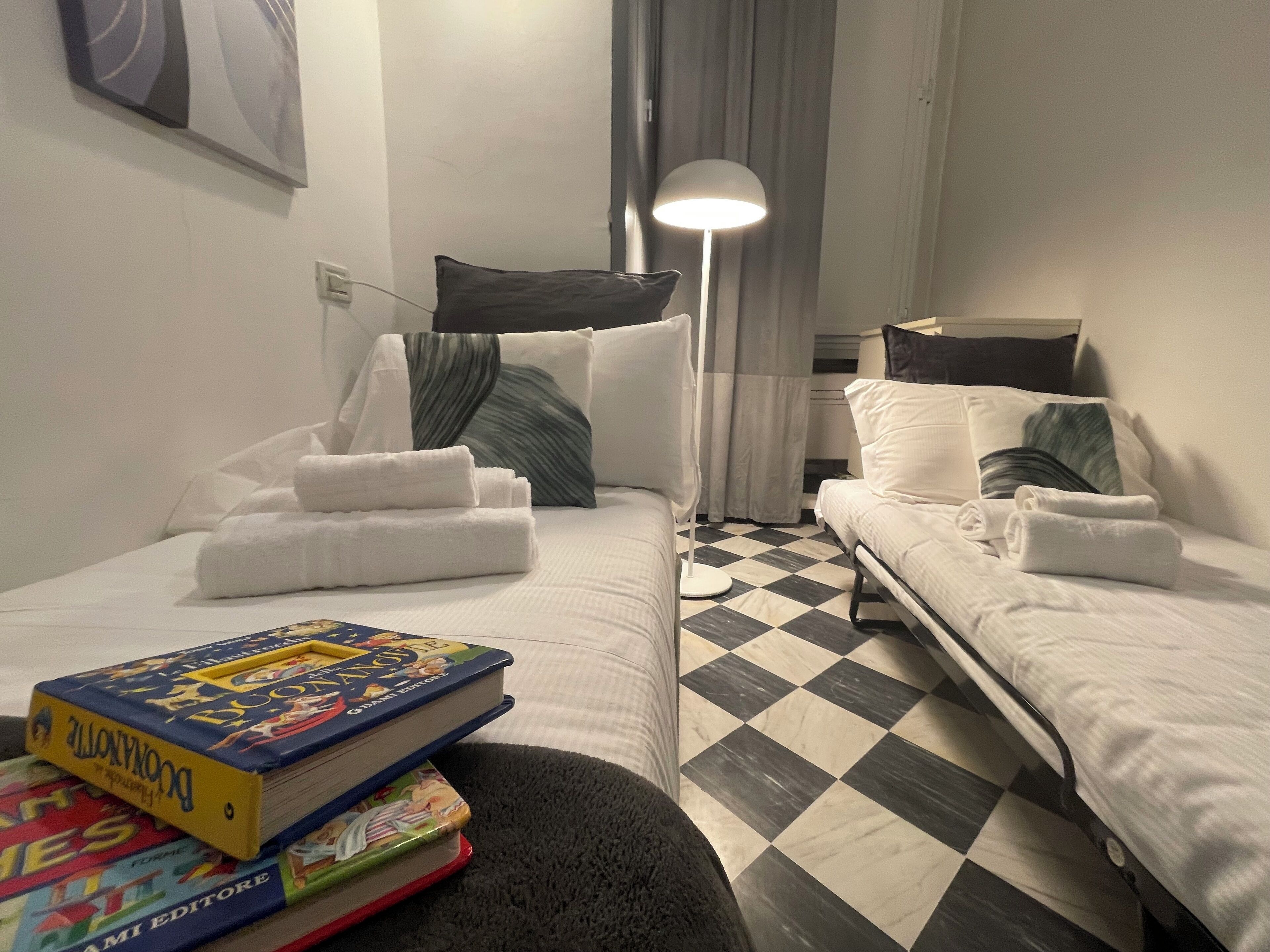 Une chambre d’hôtel avec deux lits, un sol à carreaux, une lampe et une pile de livres.