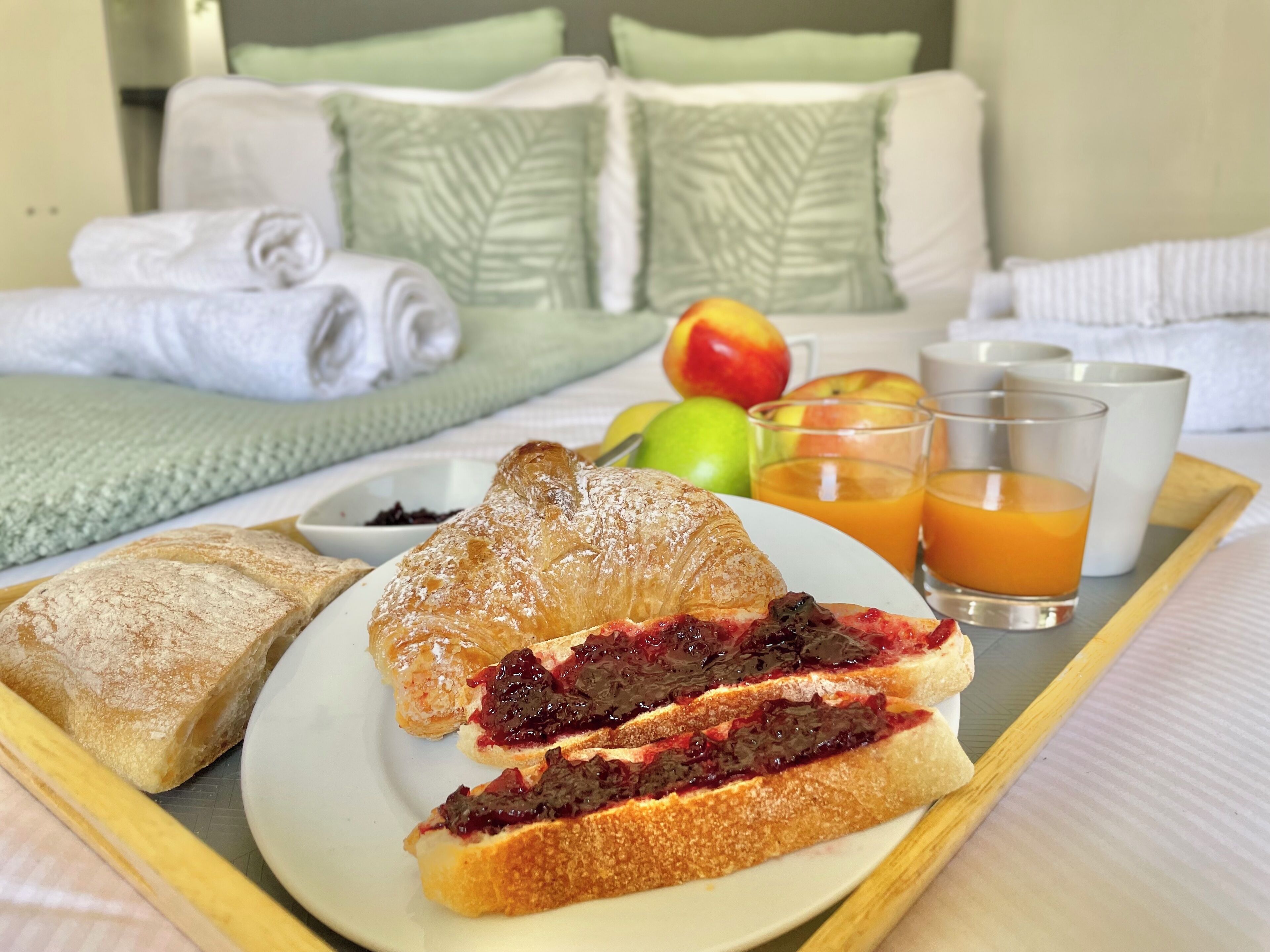 Bandeja de desayuno con cruasanes, pan con mermelada, fruta fresca y jugo de naranja sobre una cama de hotel.