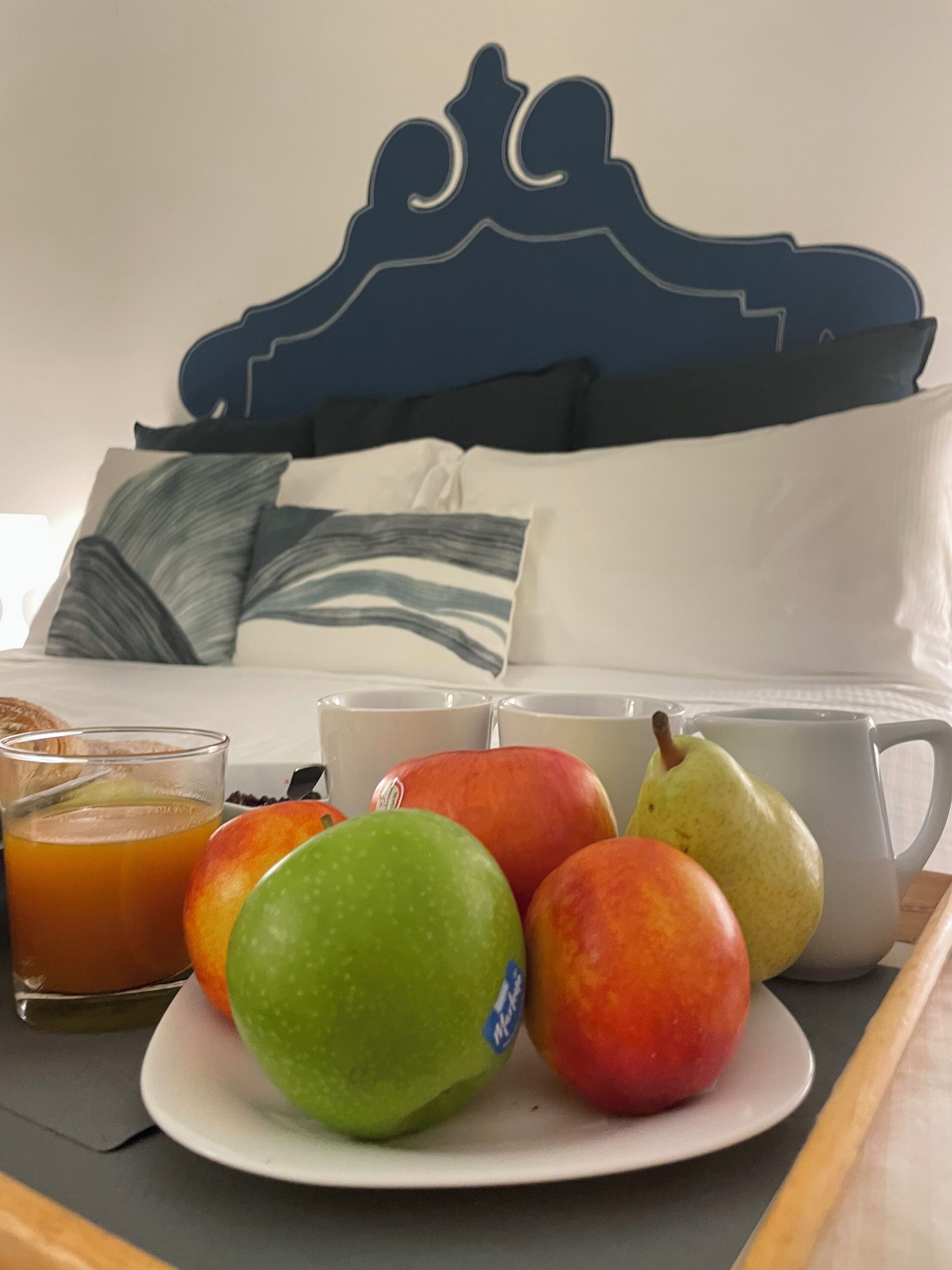 A breakfast tray with fruits and a glass of orange juice on a table, with a bed and pillows in the background.