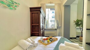 A hotel room with a bed, a tray of fruits and a cup, towels, a window with a view of buildings, and a plant. - Happy Family Apartments Cathedral (Genoa)