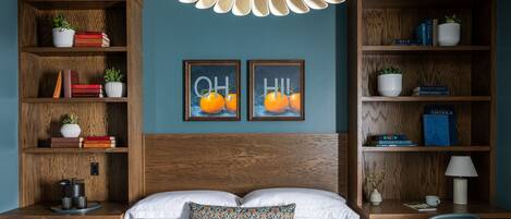 A bedroom with a wooden headboard, a bed with white linens, two framed pictures on the wall, and a desk with a chair.