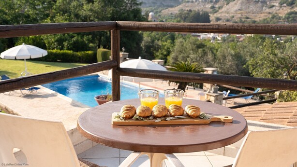 A table with croissants and orange juice on a balcony overlooking a swimming pool and lush greenery. - Villa Louna, Letymbou -  a private villa that sleeps 8 guests in 4 bedrooms (Letymbou)