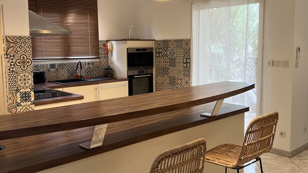 A modern kitchen with a central island, pendant lights, and a dining area with wicker chairs. - Villa Sete les Pieds Dans Leau (Sète)