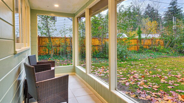 Une terrasse couverte, dotée de chaises en osier, de grandes fenêtres et offrant une vue sur un jardin avec une clôture en bois et des arbres.