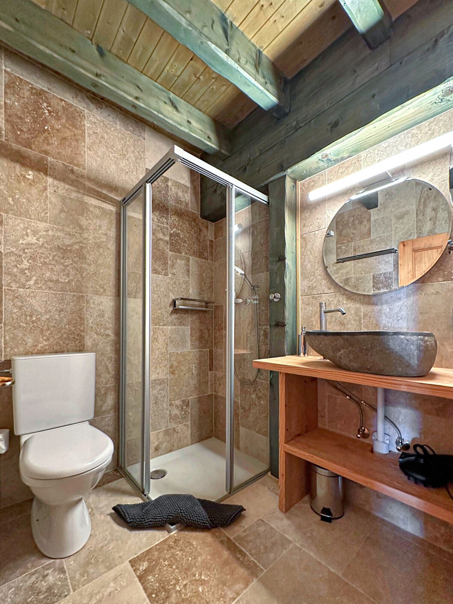 Une salle de bain avec une douche à parois de verre, des toilettes blanches, un meuble-lavabo en bois doté d’un grand évier et un miroir.