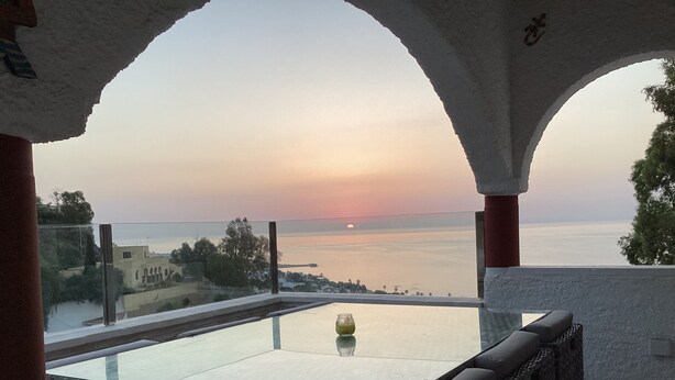 Un balcón con mesa de vidrio y mobiliario de mimbre, con vista a la puesta de sol sobre el mar.