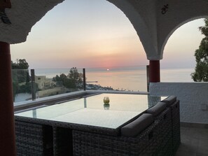 Een balkon met een glazen tafel en rieten meubilair, met uitzicht op een zonsondergang boven de zee.
