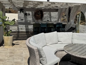 A curved wicker sofa and table in an outdoor patio area with a cactus plant and a stone wall.