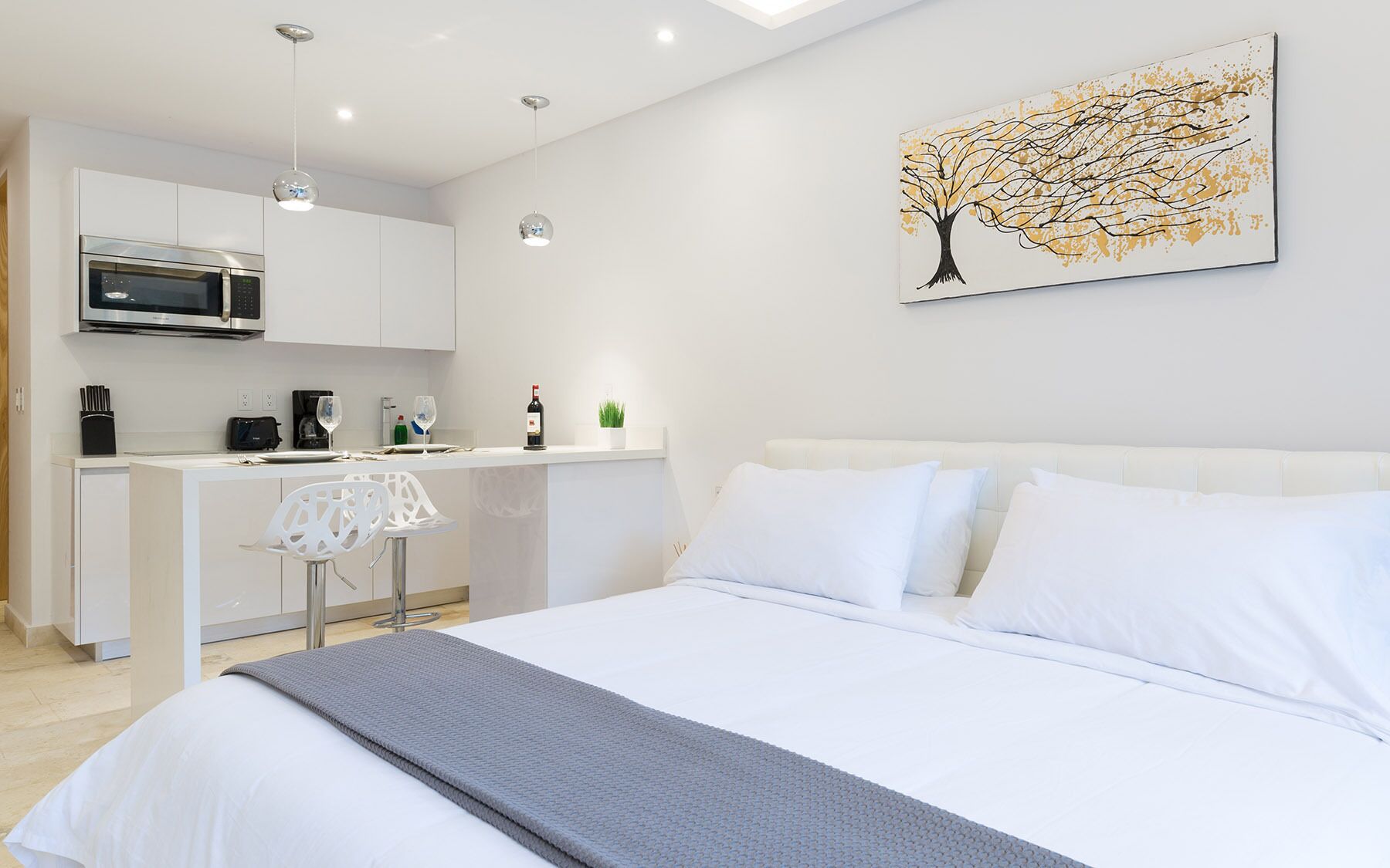 A modern bedroom with a bed, white pillows, and a gray throw blanket. Adjacent is a kitchenette with a microwave, toaster, and wine glass. A small plant is on the counter, and a painting of a tree hangs on the wall.