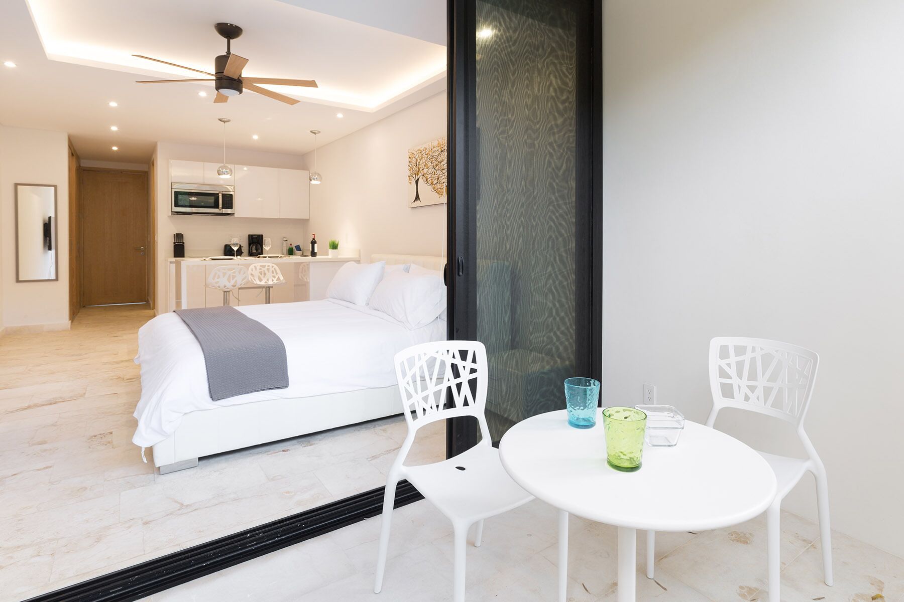 Ein modernes Hotelzimmer mit Bett, Esstisch mit Stühlen, Küchenzeile und Deckenventilator.