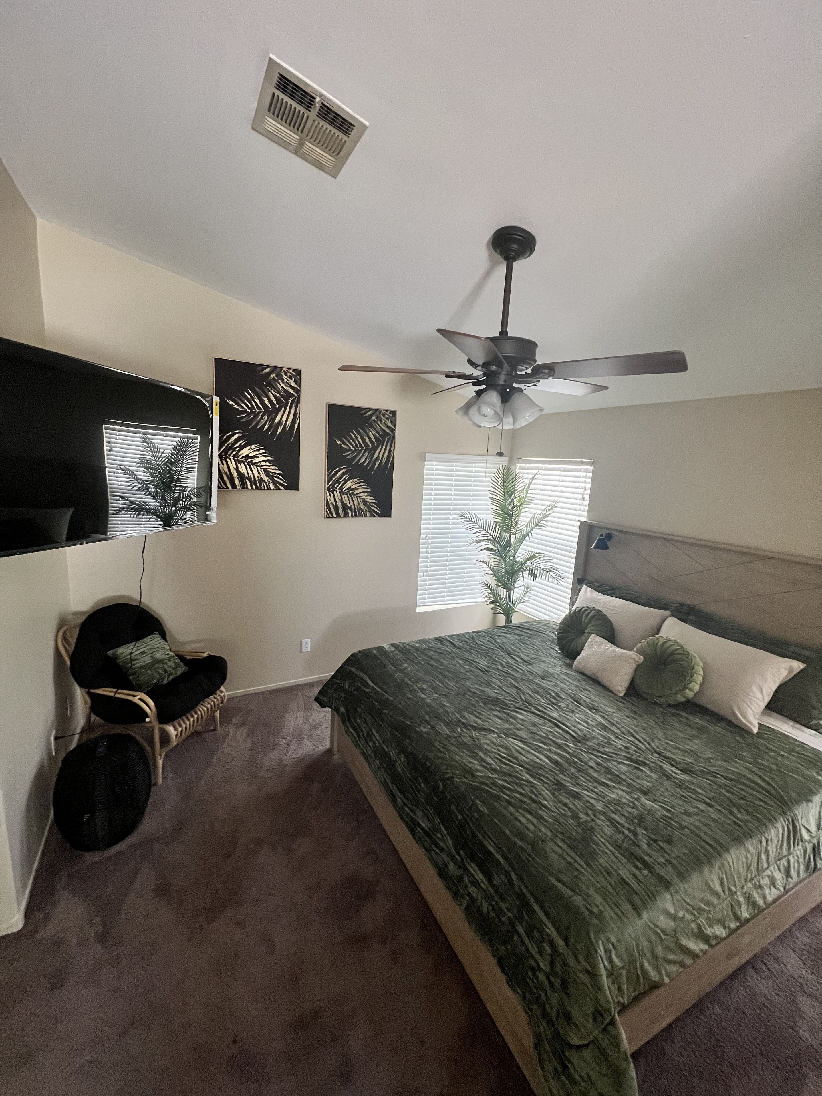 A bedroom with a bed, a ceiling fan, a window with blinds, a wall-mounted TV, and a chair.