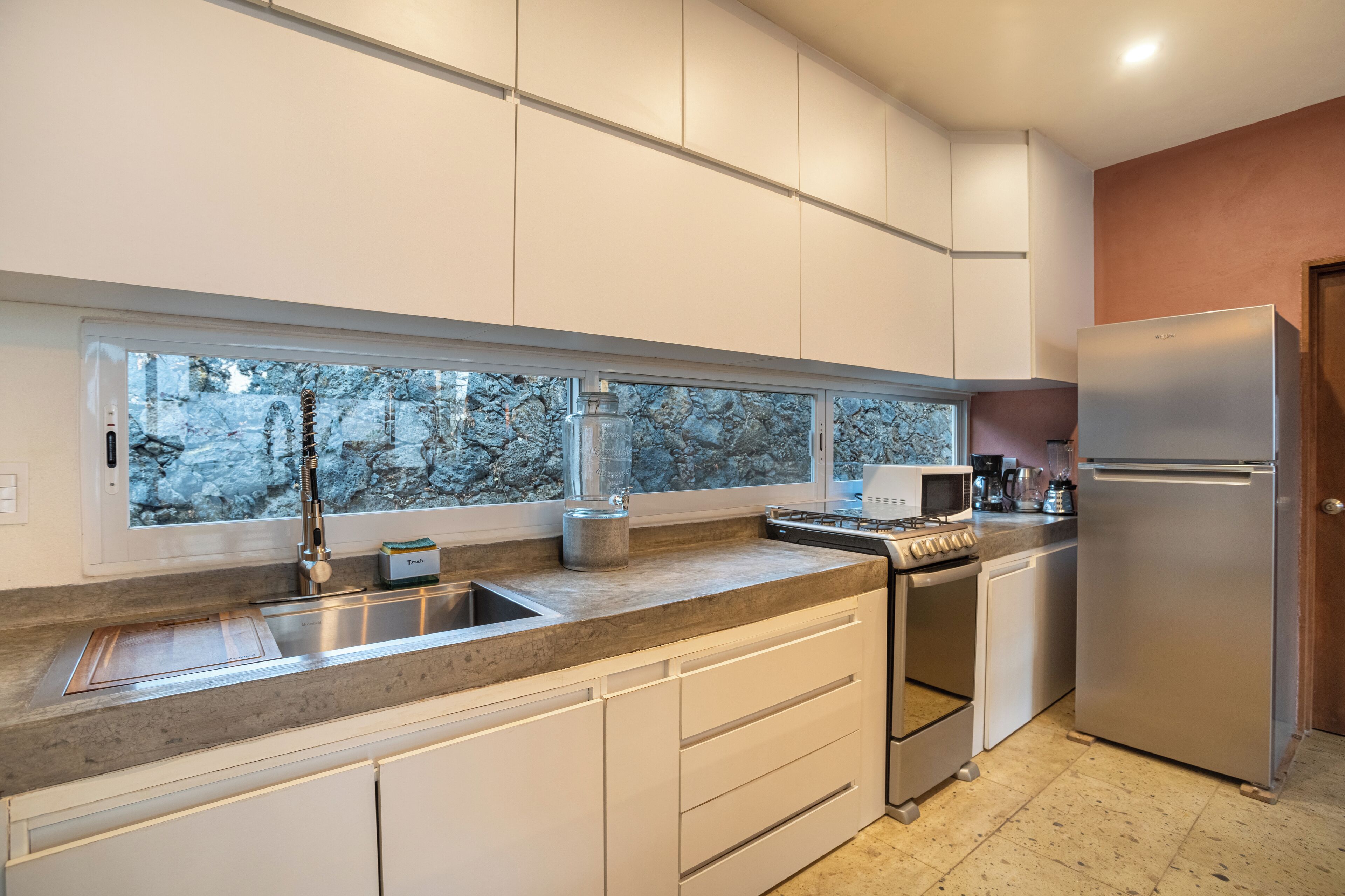 A modern kitchen with white cabinets, a stainless steel refrigerator, a built-in oven, and a window with a view of trees.