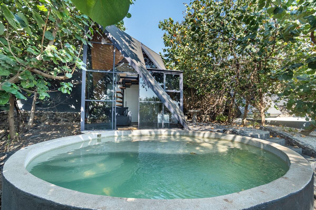 Un área de piscina exterior moderna con una estructura de cristal, rodeada de árboles y un cielo despejado.