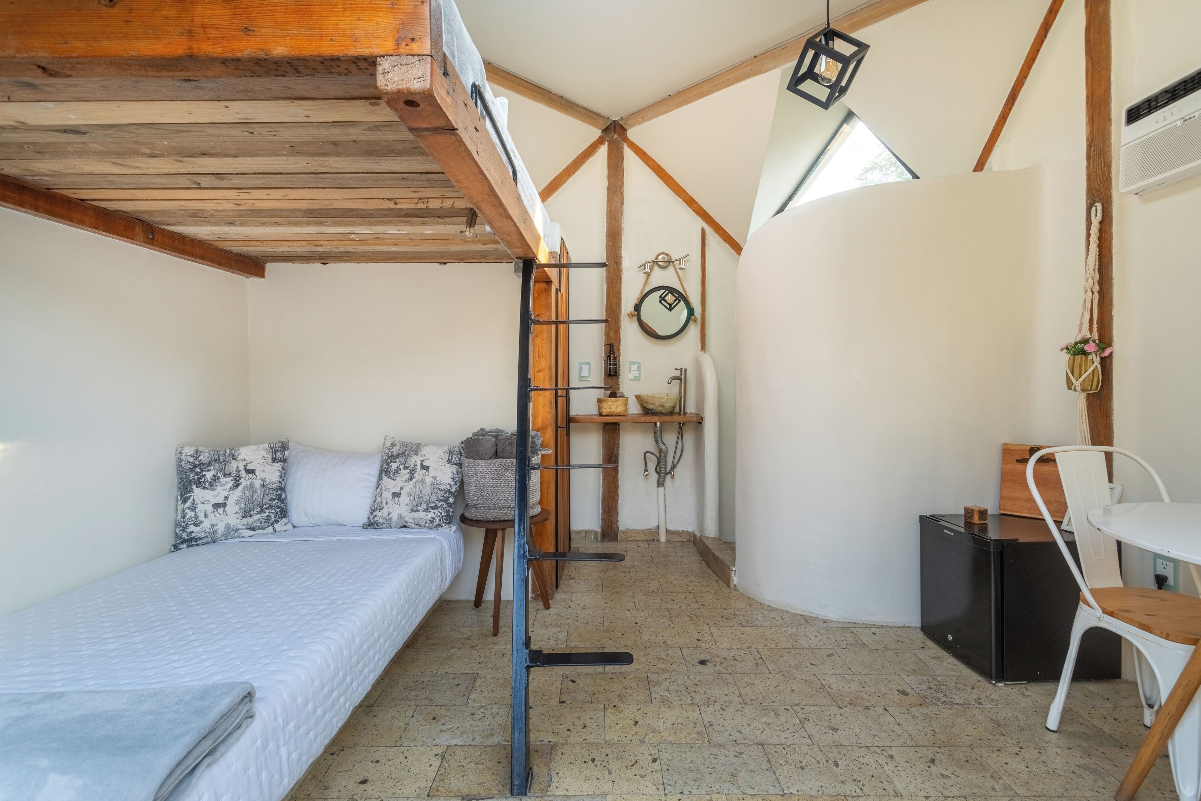 Quarto pequeno com cama, uma mesinha e uma cadeira. Uma escada encosta na parede e há uma prateleira com objetos.