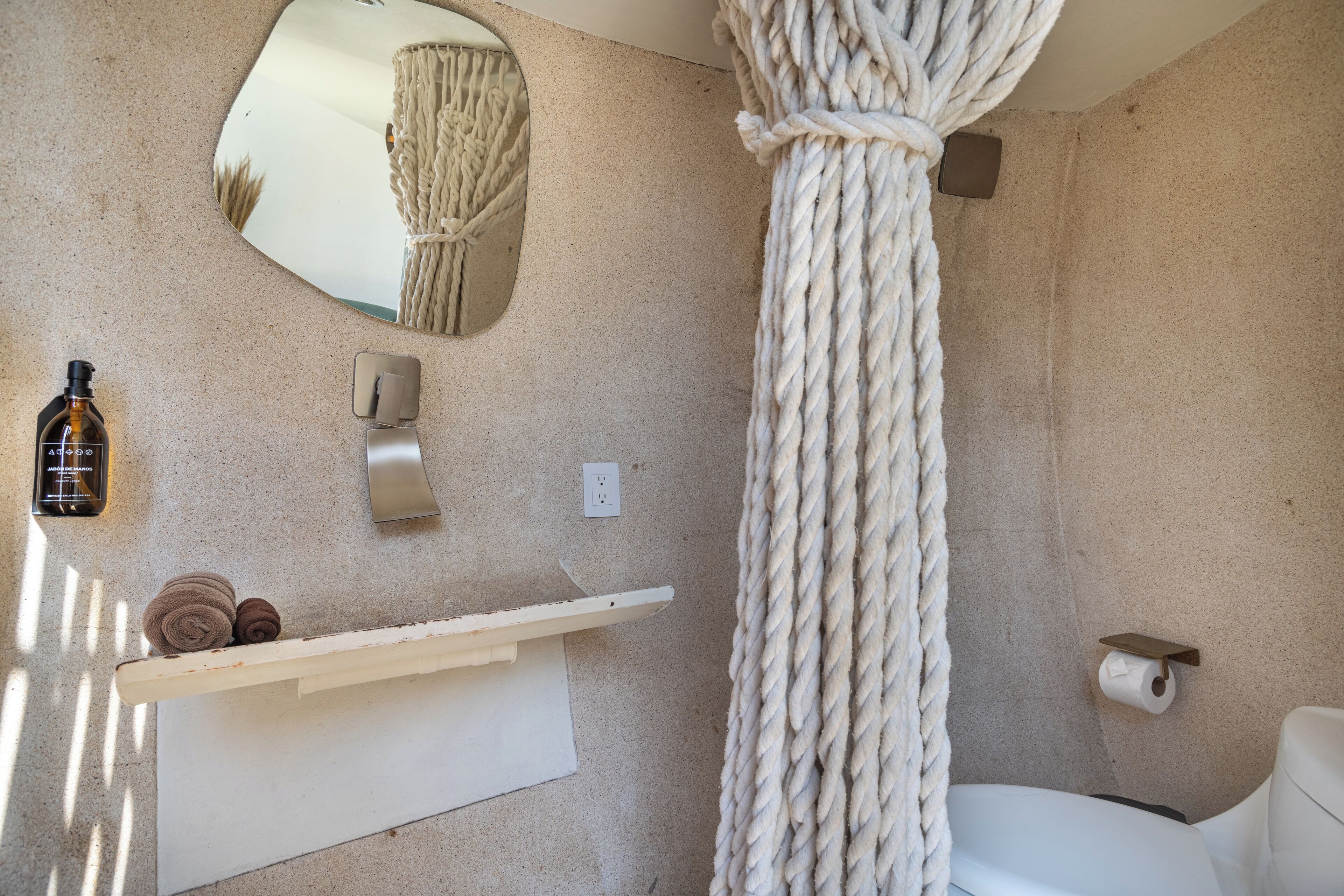 A bathroom with a rope hanging from the ceiling, a towel rack, a mirror, and a toilet paper holder.