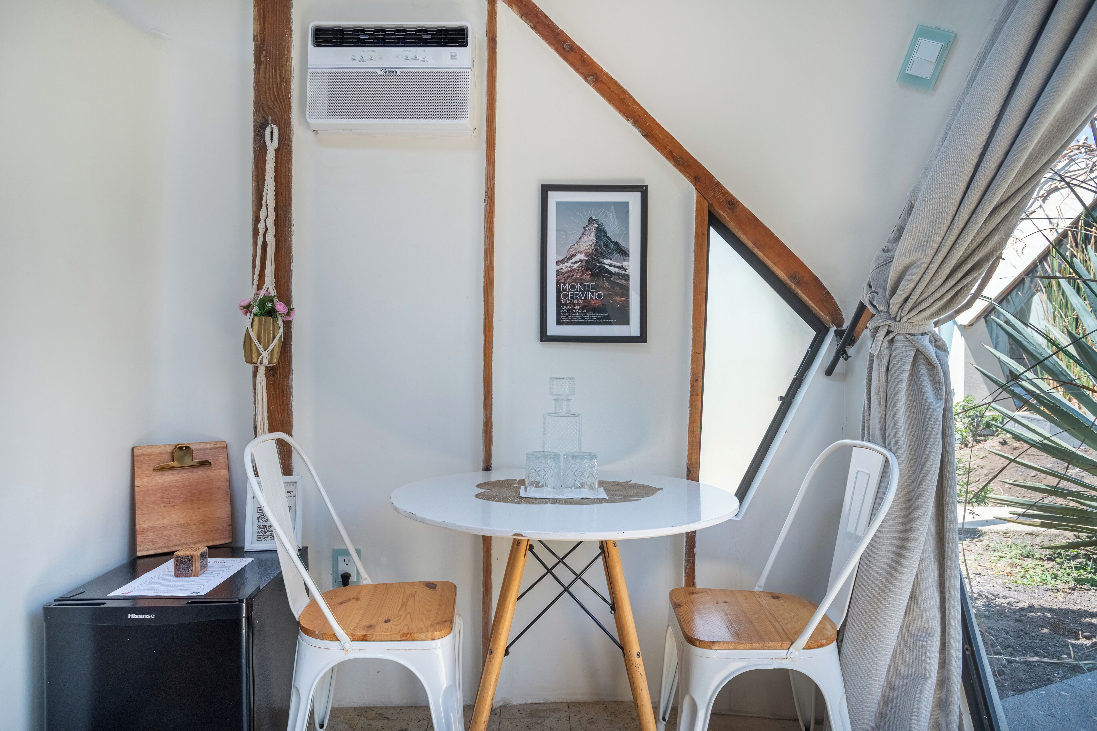 Una habitación compacta con una pequeña mesa, dos sillas, un refrigerador, un aire acondicionado de pared, un cuadro enmarcado y una cortina.
