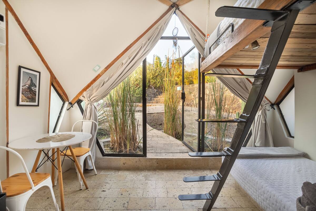 Un acogedor cuarto con forma de tienda triangular, con una mesa, sillas, una cama y vistas a un jardín.